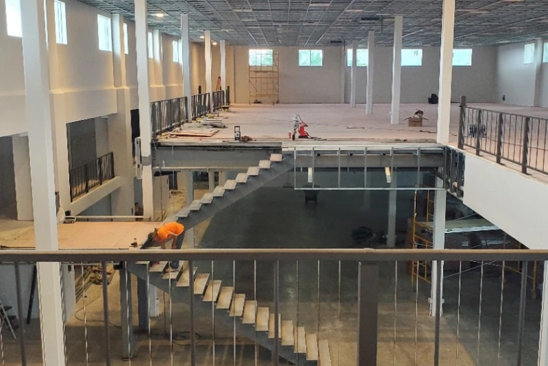 An interior shot of a large, open commercial space under construction. The photo shows an unfinished second-story level with a metal staircase, and a worker on the lower level. The space has tall white pillars and large windows, suggesting an office or retail buildout.